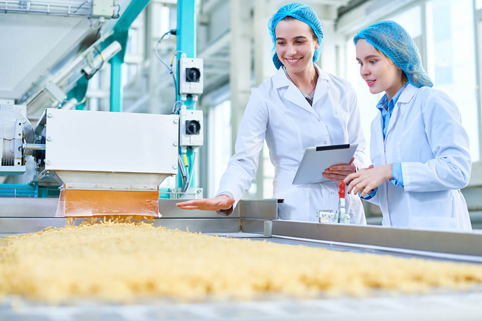 Due donne in camice da laboratorio ispezionano una linea di produzione con materiale sfuso.