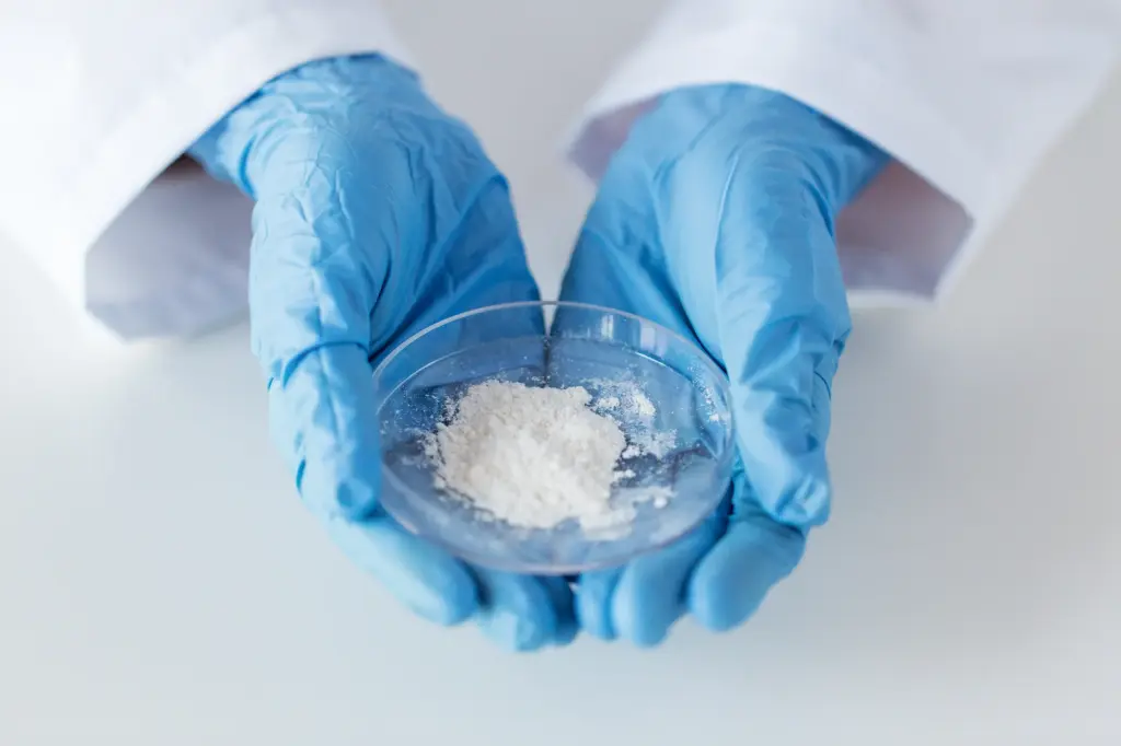 Inquadratura ravvicinata di una capsula di Petri contenente una polvere bianca, tenuta da due mani che indossano guanti protettivi – come immagine simbolica per i prodotti chimici