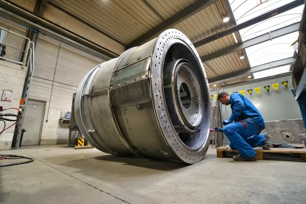SPALECK, un operaio meccanico, è inginocchiato davanti a un grande gruppo saldato e sta montando dei componenti. Il sottogruppo fa parte di un grande sottogruppo saldato destinato a una turbina a gas. L'immagine mostra la produzione presso SPALECK.