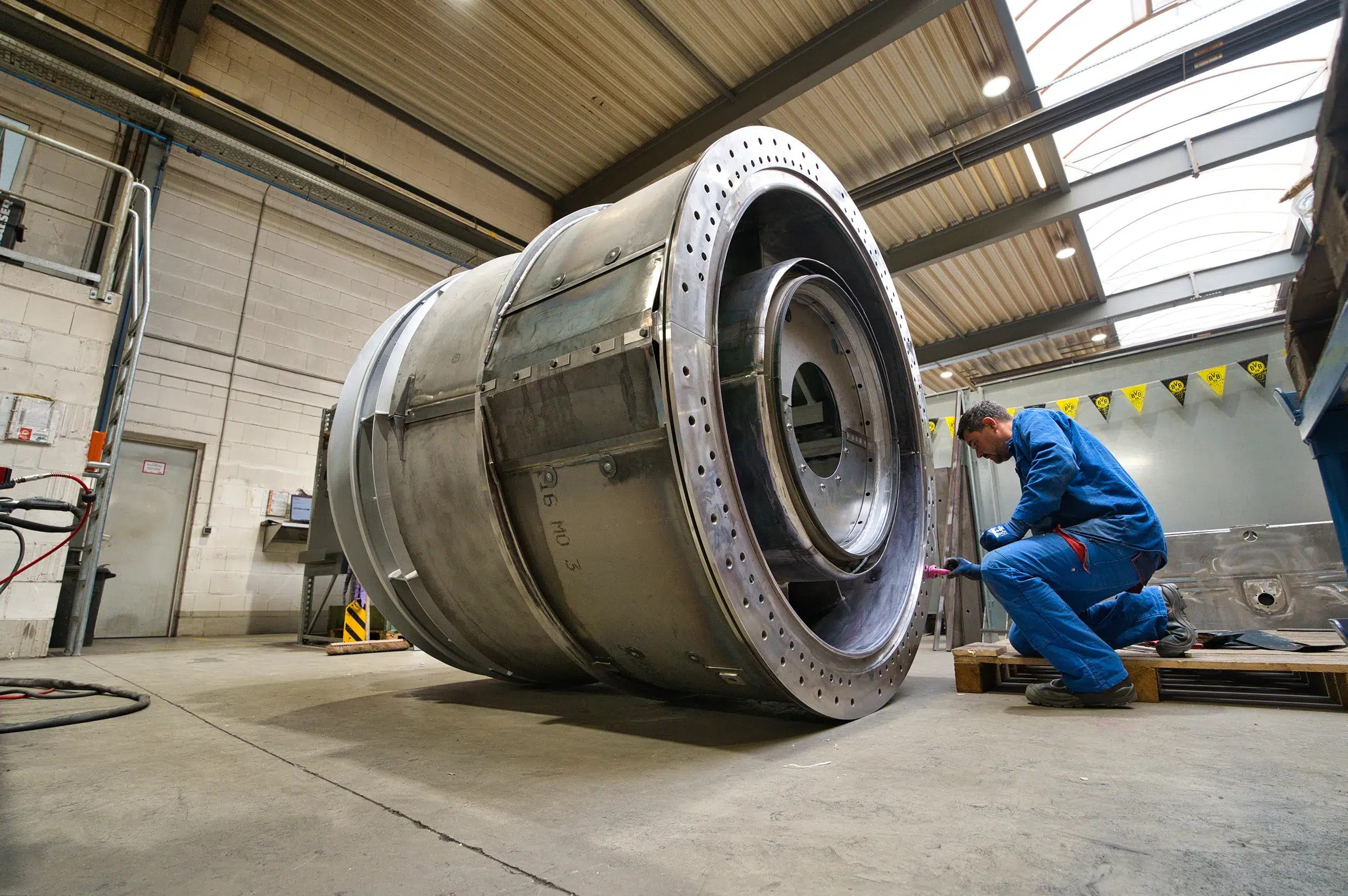 SPALECK, un operaio meccanico, è inginocchiato davanti a un grande gruppo saldato e sta montando dei componenti. Il sottogruppo fa parte di un grande sottogruppo saldato destinato a una turbina a gas. L'immagine mostra la produzione presso SPALECK.