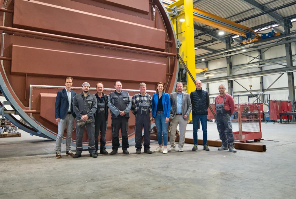 Un gruppo di persone è in piedi davanti a un grande macchinario industriale in un capannone industriale.