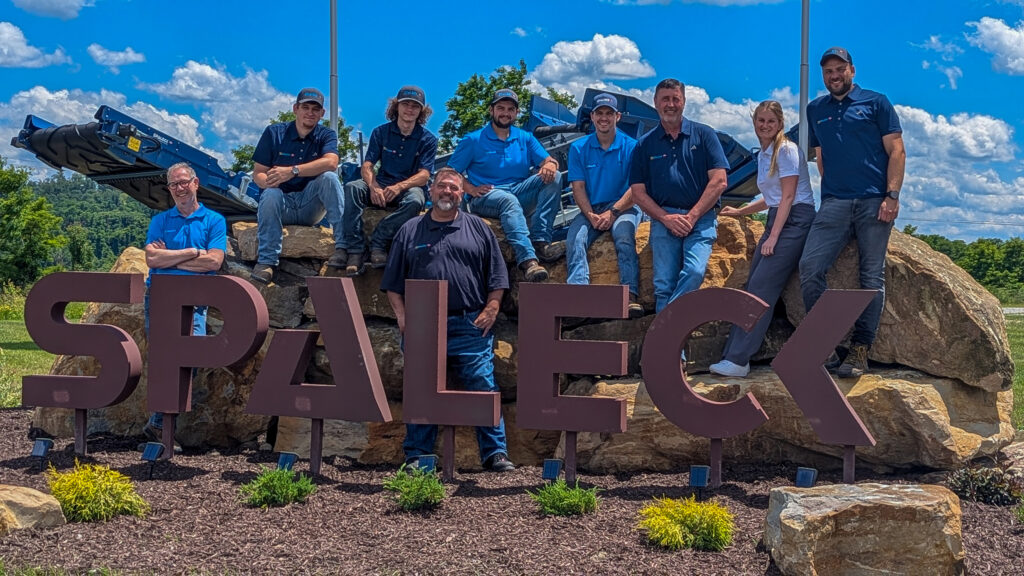 Foto di gruppo dello Spaleck USA davanti al logo Spaleck a Charleroi in occasione dell'inaugurazione della sede, sotto un cielo azzurro