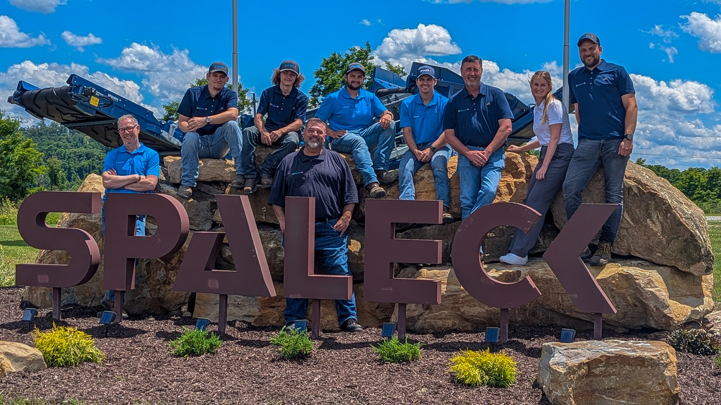 Foto di gruppo dello Spaleck USA davanti al logo Spaleck a Charleroi in occasione dell'inaugurazione della sede, sotto un cielo azzurro